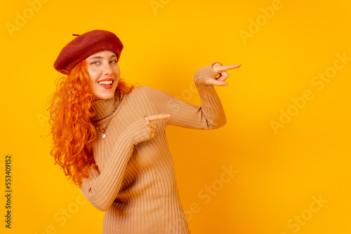 Red-haired woman in a red beret on a yellow background, studio shot, pointing at copy space, smiling