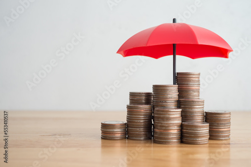 Stacked coins is protected by red umbrella on wooden table white wall background copy space. Assets wealth, money saving or money investment protection, security by insurance concept. Risk management.