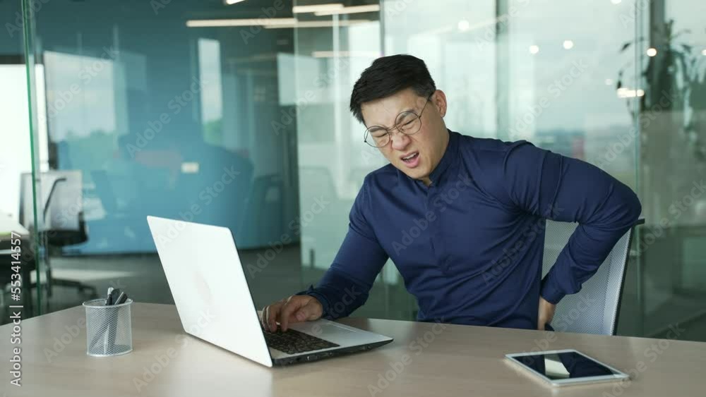 Overworked tired asian businessman IT programmer with severe back pain after hard work on computer at desk in the modern workplace Entrepreneur with glasses suffers from tension in the spine indoors