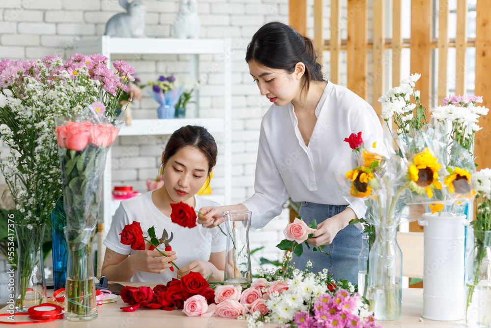 Millennial Asian young beautiful professional female flower shopkeeper decorator florist worker smiling holding flower bunch bouquet colleague arranging decorating stalk in floral store.
