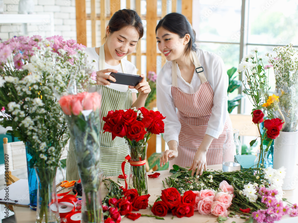Millennial two Asian young professional female flower shopkeeper owner decorator wearing apron standing smiling using smartphone taking photo of decorating red roses bunch bouquet in vase in store