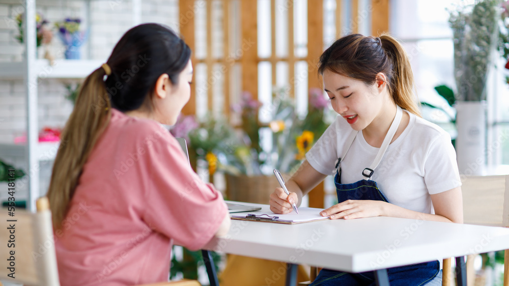 Millennial Asian young professional female shopkeeper decorator florist owner worker wearing jeans apron sitting smiling using pen writing customer need on paper clipboard on table in flowers store