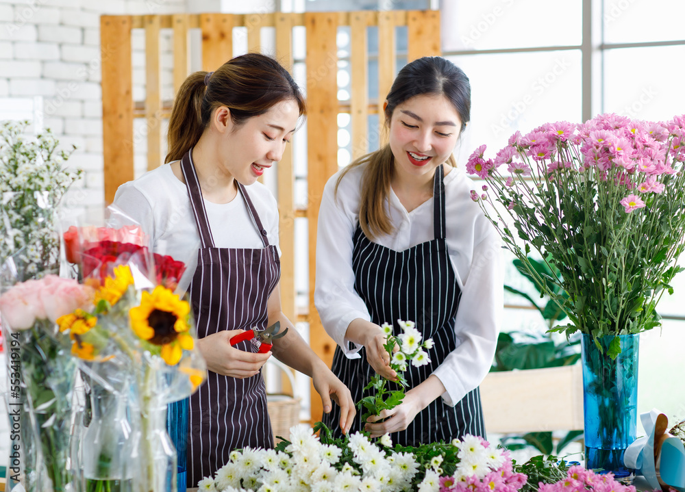 Millennial Asian young female flower shopkeeper decorator florist worker in apron smiling holding white flower bunch bouquet colleague use scissors cutting arranging decorating stalk in floral store