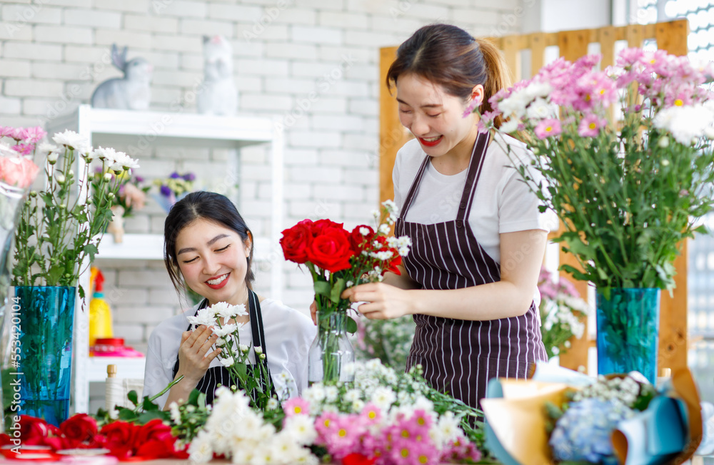 Millennial Asian young female flower shopkeeper decorator florist worker in apron smiling holding flower helping bunch bouquet colleague arranging decorating stalk in floral store.