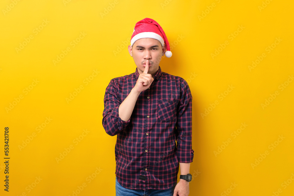 Young Asian man in Santa hat showing silence gesture, holding finger near lips over yellow studio background. Happy New Year 2023 celebration merry holiday concept