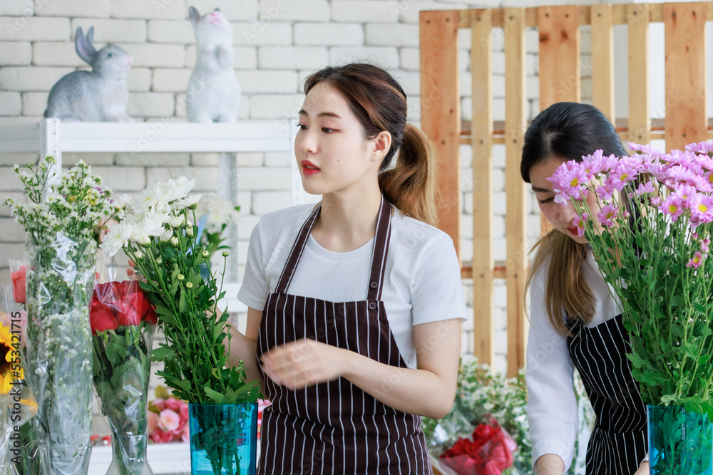 Millennial Asian young female flower shopkeeper decorator florist worker in apron smiling holding white flower bunch bouquet colleague use scissors cutting arranging decorating stalk in floral store
