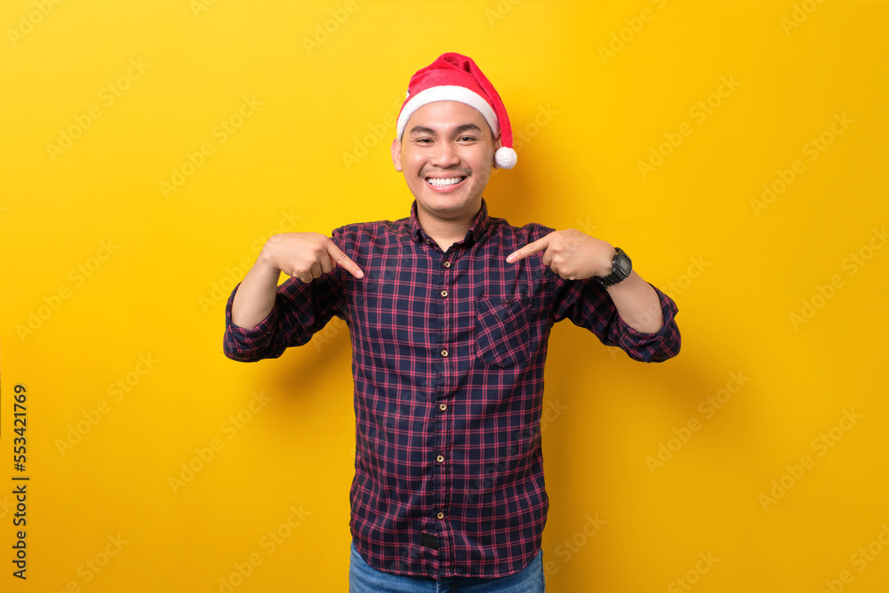 Cheerful young Asian man in Santa hat pointing fingers down and advertising something over yellow studio background. Happy New Year 2023 celebration merry holiday concept