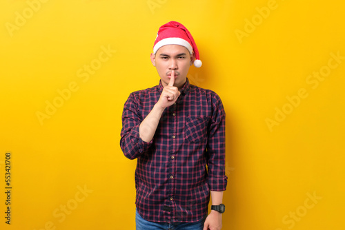 Young Asian man in Santa hat showing silence gesture, holding finger near lips over yellow studio background. Happy New Year 2023 celebration merry holiday concept