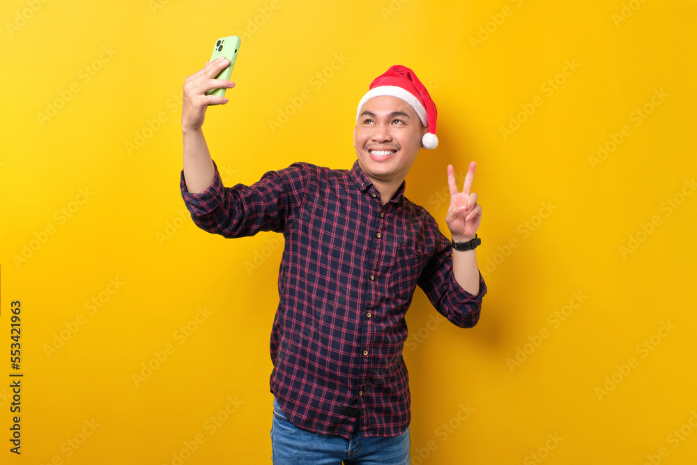 Cheerful young Asian man in Santa hat taking selfie on smartphone, showing victory sign over yellow studio background. Happy New Year 2023 celebration merry holiday concept