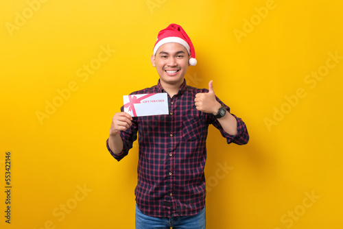 Smiling young Asian man in Santa hat holding gift certificate, showing thumb up over yellow studio background. Happy New Year 2023 celebration merry holiday concept