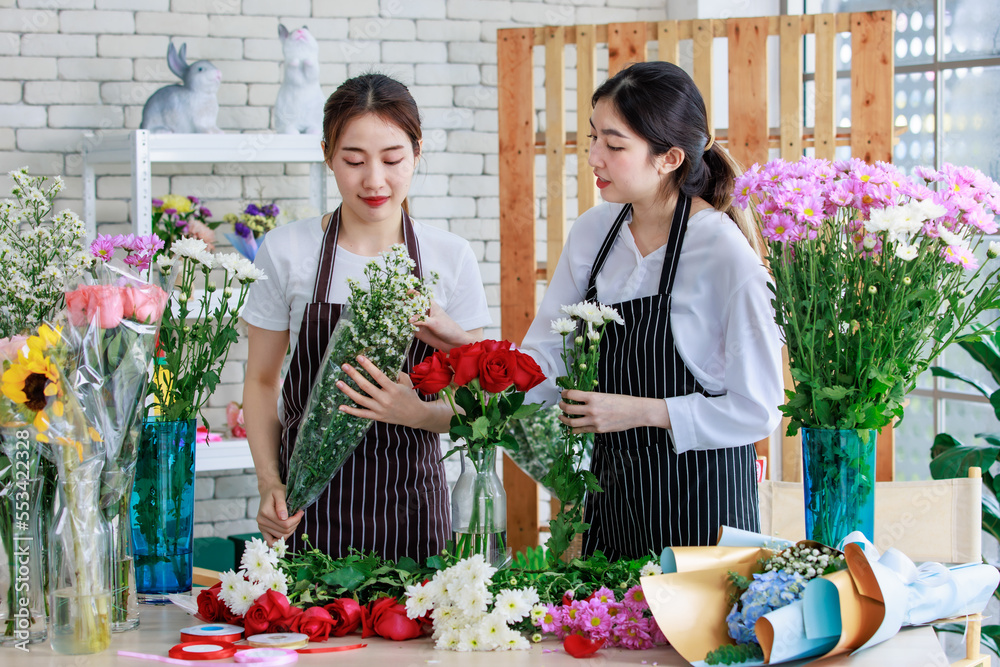 Millennial Asian young female flower shopkeeper decorator florist worker in apron smiling holding flower bunch bouquet colleague arranging decorating stalk in floral store.