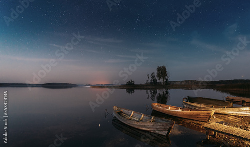 Moored Old Shabby Wooden Fishing Rowboats   Left Afloat On The Motionless Clear River Lake Water Next To Waterside. Amazing Glowing Stars Effects Above Lake. Night Starry Sky Soft Colors.