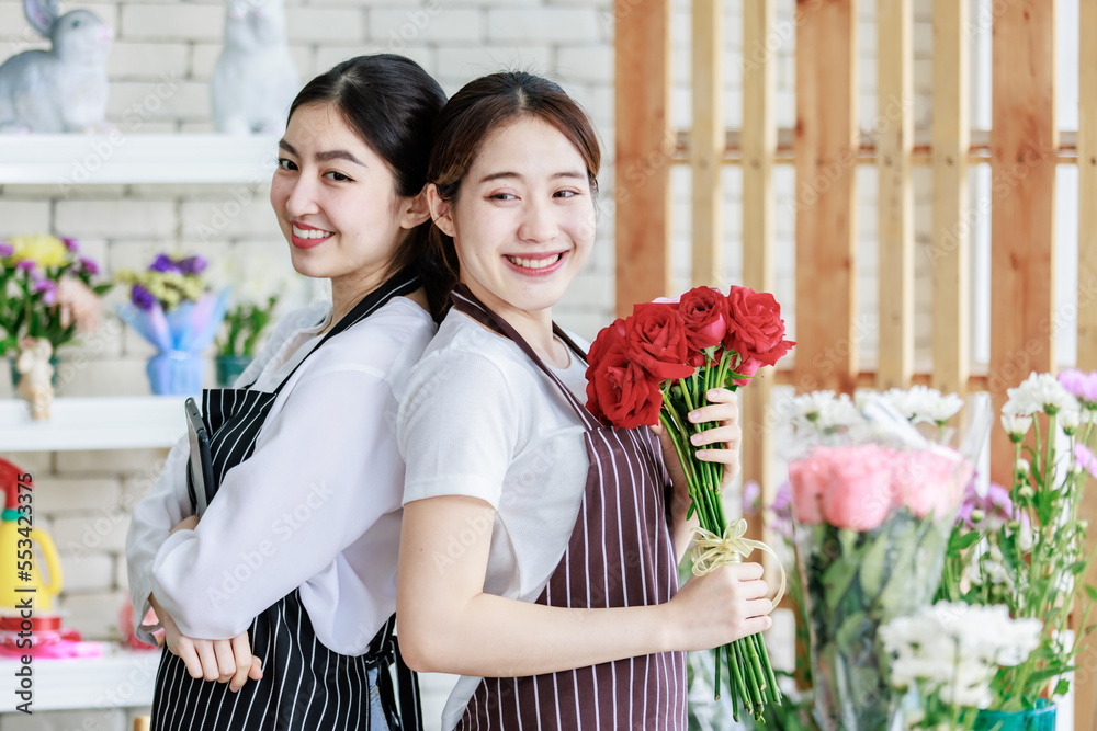 Millennial Asian young professional flower shopkeeper florist employee worker wearing apron standing smiling hold red roses bunch bouquet leaning back with colleague posing take photo in floral store