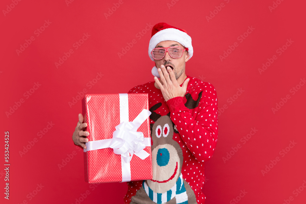 surprised man in winter sweater and santa claus hat. xmas guy with present box on red background. surprised man in winter sweater and santa claus hat. xmas guy with present box on red background.