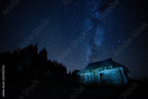 Night photo of an old wooden house in the mountains among the fir trees and the milky way.