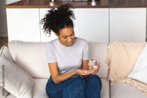 Young beautiful smiling African American woman holding painkiller, antidepressant pill and glass of water sitting on the couch at home photo