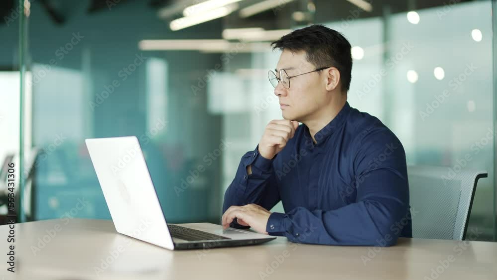 Busy middle aged IT specialist programmer with glasses working typing coding on laptop computer in modern office Asian entrepreneur professional developer thinking analyses finance digital data indoor