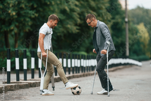 Having fun, playing soccer. Two men with crutches is outdoors on the road