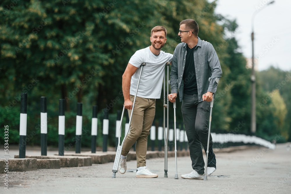 Standing and smiling. Two men with crutches is outdoors on the road Standing and smiling. Two men with crutches is outdoors on the road