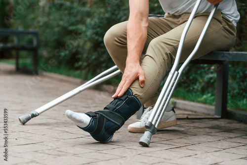 Touching the leg. Man with crutches is in the park outdoors