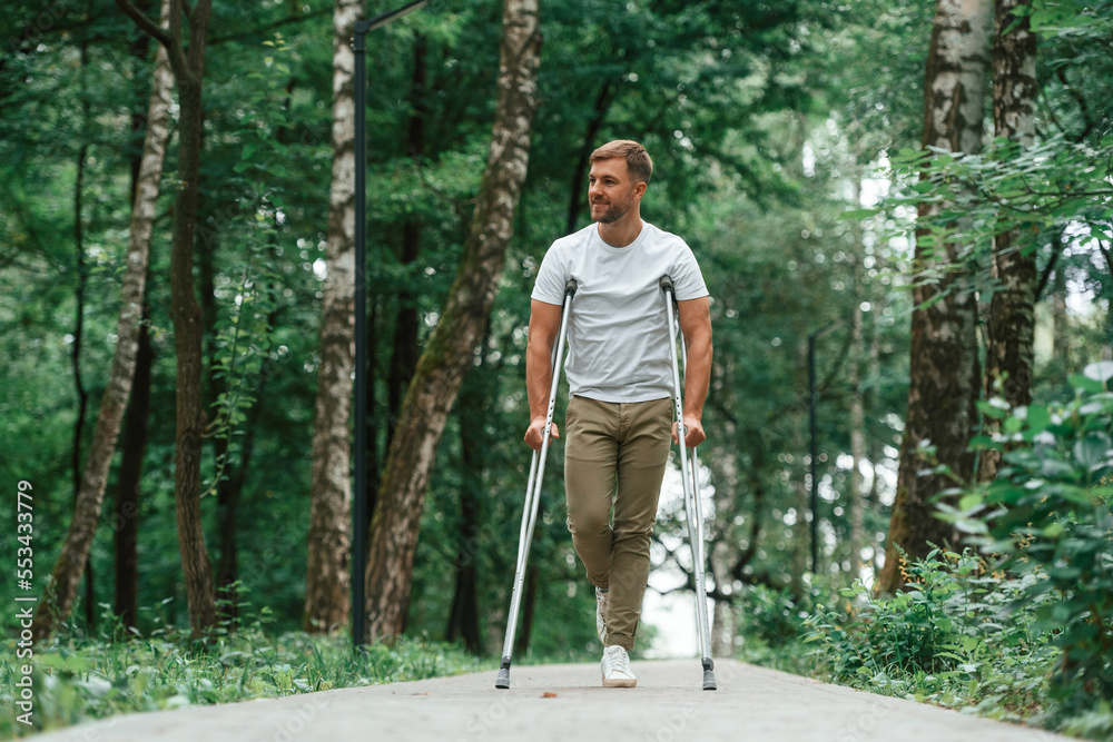 Walking in the beautiful park. Man with crutches is outdoors. Having leg injury Walking in the beautiful park. Man with crutches is outdoors. Having leg injury