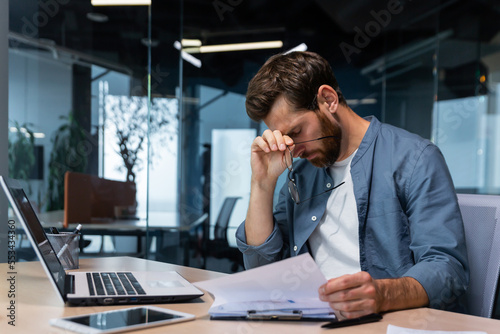 Billede på lærred Upset businessman behind paper work inside modern office, mature man with beard reading financial reports and account documents unhappy with results and disappointed with achievements
