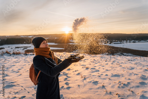 A woman in a snow-covered field tosses snow with her hands against the backdrop of a sunset photo