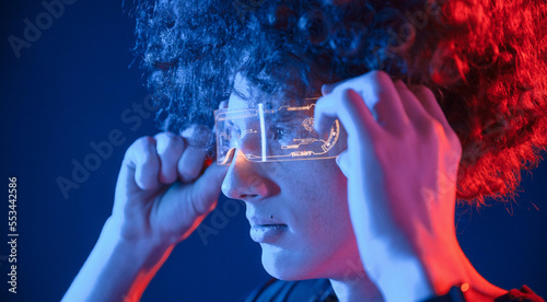 Wearing the futuristic glasses. Young man with curly hair is indoors illuminated by neon lighting