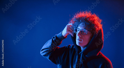 Wearing the hood. Young man with curly hair is indoors illuminated by neon lighting