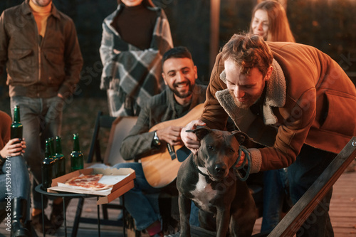 Group of people is having fun together. Man playing guitar, his friend is playing with dog