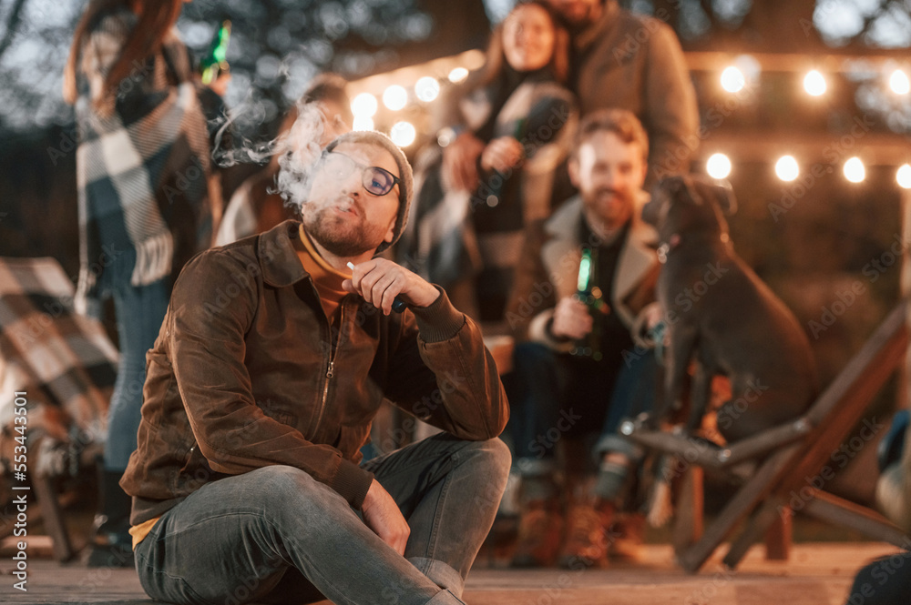 Man is smoking. Group of people is spending time together on the backyard at evening time