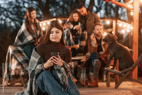 Woman with smartphone is sitting. Group of people is spending time together on the backyard at evening time