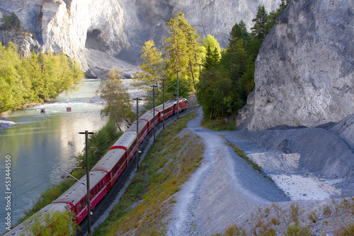 Anterior Rhine valley gorge with Rhine River and red RHB train arriving at railway station Versam-Safien on a sunny autumn morning. Photo taken September 26th, 2022, Versam Station, Switzerland.