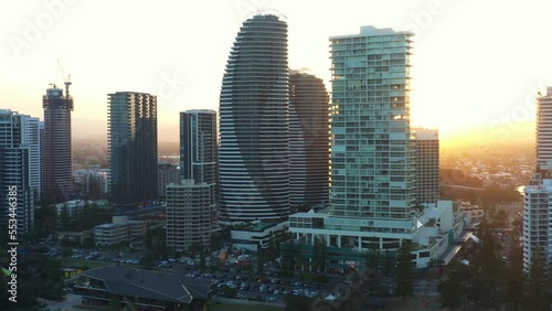 Aerial fly around broadbeach gold coast, panoramic panning view of waterfront cityscape with high rise apartments and resort hotels along the beach with sun peeking through in between buildings. photo