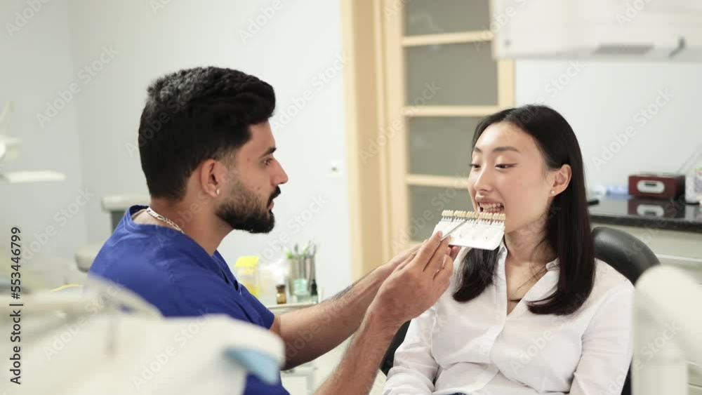 Dentist choosing color of filling from palette. Back view of male dentist checking and selecting color of young asian woman teeth indoors in modern hospital. Dentist choosing color of filling from palette. Back view of male dentist checking and selecting color of young asian woman teeth indoors in modern hospital.