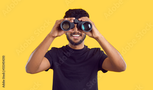 Happy ethnic man looking through binoculars looking for profitable advertising offers, isolated on yellow background. Dark-skinned bearded man in black t-shirt looking at you through binoculars
