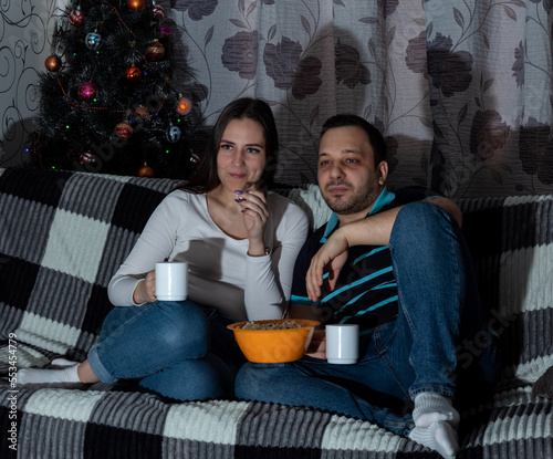 Young woman and man are sitting on couch with mugs, eating popcorn while watching TV. Close-up. Selective focus. Images for articles about movies, hobbies, leisure.