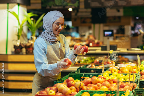 Murais de parede Female seller in hijab browsing and checking apples in supermarket, woman in apron smiling at work in store in fruit and vegetable department