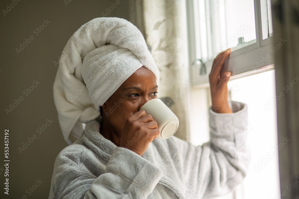 Woman in bathrobe and towel turban looking through window and drinking from mug