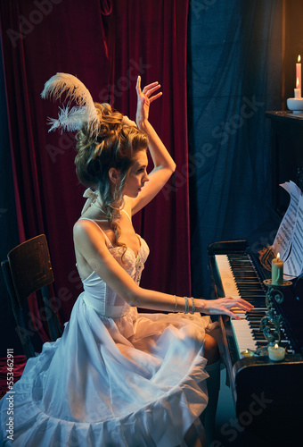 Elegance and tenderness. Portrait of beautiful young woman in image of medieval person in white dress sitting at piano and playing. Comparison of eras, beauty, history, art