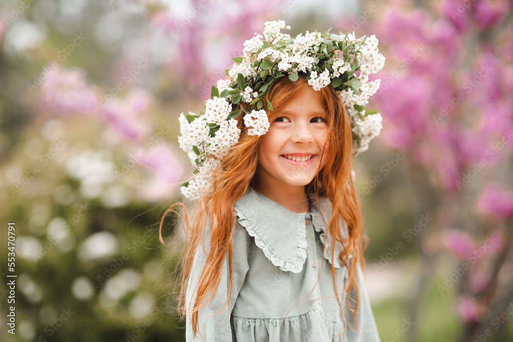 Fototapeta premium Cute laughing little kid girl 3-4 year old with long curly red hair wear floral wreath and stylish rustic dress over nature background in garden outdoor. Springtime.