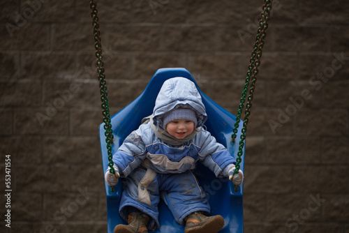 chubby Kid with ruddy cheeks rides on a swing in a blue overalls with a hood