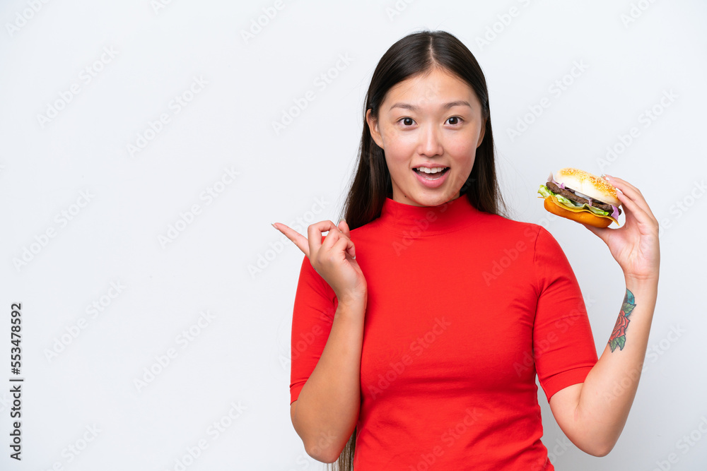 Young Asian woman holding a burger isolated on white background surprised and pointing finger to the side Young Asian woman holding a burger isolated on white background surprised and pointing finger to the side