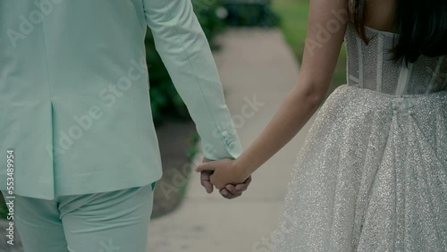 the bride and groom walking along the path in the garden hold hands. slow motion video from the back photo