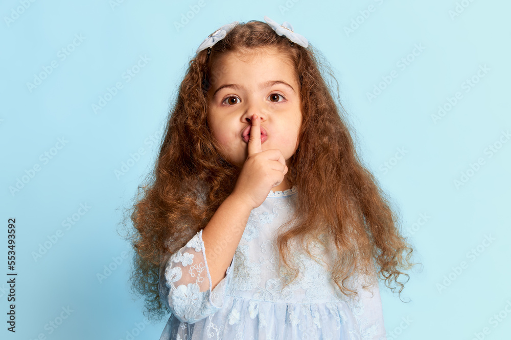 Secrets. Studio shot of preschool age girl in blue dress press her finger to her lips isolated over blue background. Happiness, kids fashion concept Secrets. Studio shot of preschool age girl in blue dress press her finger to her lips isolated over blue background. Happiness, kids fashion concept