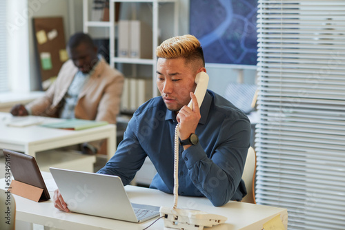 Photography Portrait of Asian man speaking on corded phone and using laptop while working in