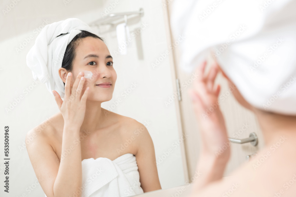 Happy young Asian woman applying face lotions while wearing a towel and touching her face in bathroom Happy young Asian woman applying face lotions while wearing a towel and touching her face in bathroom