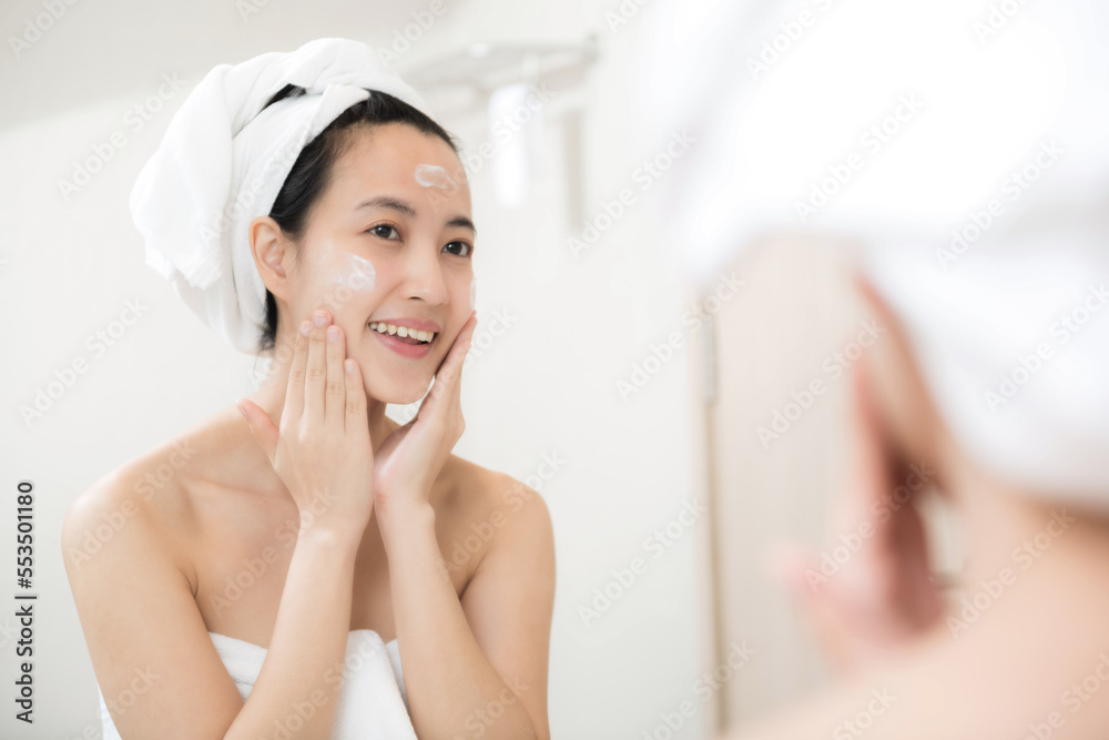 Happy young Asian woman applying face lotions while wearing a towel and touching her face in bathroom Happy young Asian woman applying face lotions while wearing a towel and touching her face in bathroom