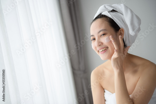 Happy young Asian woman applying face lotions while wearing a towel and touching her face. Daily makeup and skincare