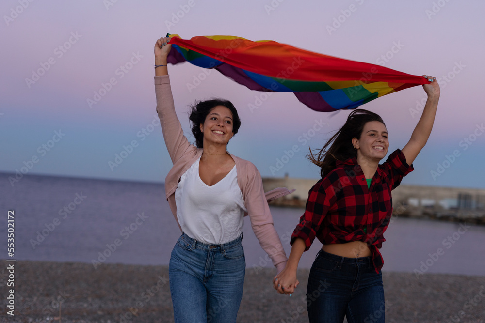Beautiful lesbian young couple embraces and holds a rainbow flag. Girls enjoy at the beach